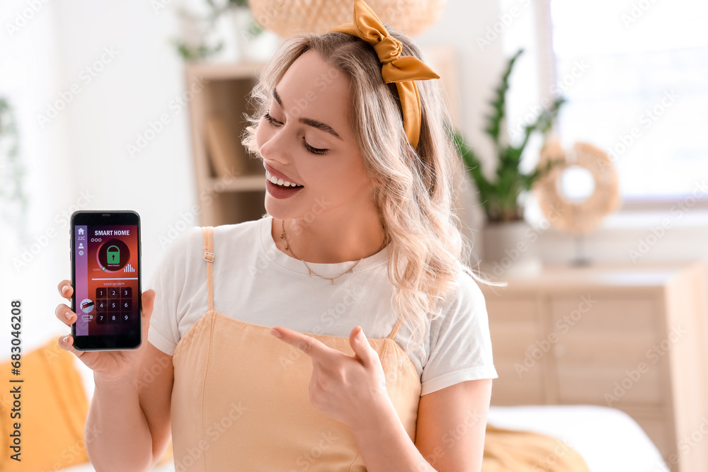 Young woman pointing at mobile phone with smart home security system control panel in bedroom