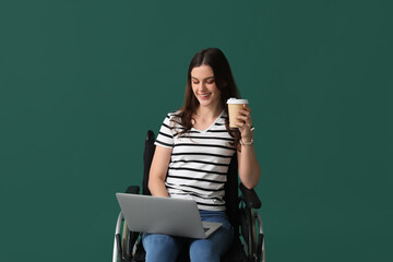 Wall Mural - Young woman with cup of coffee using laptop in wheelchair on green background