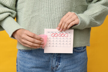 Canvas Print - Young woman holding menstrual cup and calendar with marked days of menstruation on yellow background