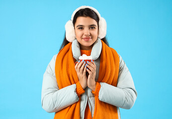 Wall Mural - Young woman in winter clothes holding cup of cocoa with marshmallows on blue background