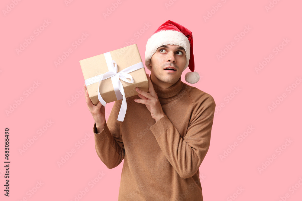 Handsome young man in Santa hat shaking Christmas gift on pink background