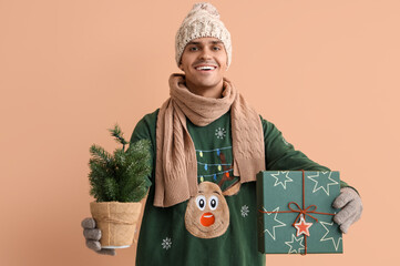 Canvas Print - Young man in Christmas sweater with fir tree and gift on beige background