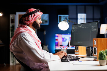 Image shows an Arab software developer working at the pc monitor, seated at a workstation processing algorithm. Muslim Coder uses a desktop computer to work on the user interface.