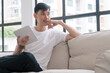 © kaew6566 - Handsome asian casual man holding digital tablet looking window thinking hand under chin sitting on sofa relax in morning apartment. Healthy guy thoughtful using tablet online. Lifestyle technology.