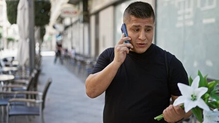 Canvas Print - Young latin man, confidently talking on his phone, grinning widely as he holds a bouquet of flowers, standing casually on a city street.