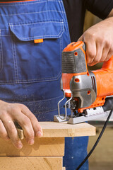 Wall Mural - Close up of experienced carpenter in work clothes and small buiness owner  carpenter saw and processes the edges of a wooden bar with a jig saw  in a workshop