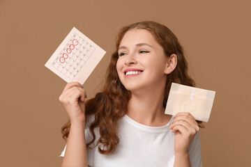 Wall Mural - Young woman holding pad and calendar with marked days of menstruation on brown background