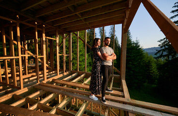 Man and woman inspecting future wooden frame dwelling nestled in the mountains near forest. Youthful couple at construction site in early morning. Concept of contemporary ecological construction.