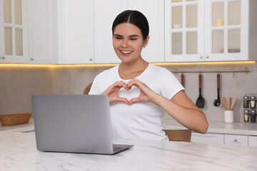 Wall Mural - Happy young woman having video chat via laptop and making heart at table in kitchen