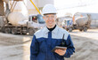 Modern technologies industrial cement plant concept. Engineer in uniform and hard hat uses tablet computer to control equipment trucks at production site