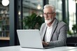 © photolas - Old businessman sitting in front of a laptop in a modern office, coworking space. A gray-haired serious man works alone.