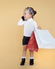 Wall Mural - Cute little girl holding a package in hand and talking on a phone, dressed in a casual clothes, posing in studio, isolated beige background.