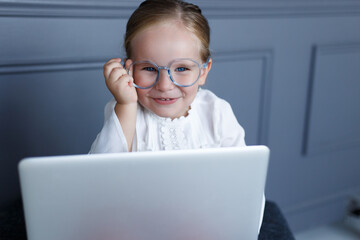 Wall Mural - Front image of a pretty smiling little child in eyeglasses, working on laptop.