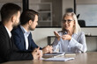 © fizkes - Positive senior boss woman in stylish glasses speaking to younger professional men, sitting at meeting table, explaining work strategy, smiling. Mature business coach training employees