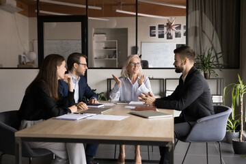 Canvas Print - Senior company owner woman talking to team of younger managers on corporate meeting, sitting at boss place at large table, explaining teamwork strategy, smiling, discussing collaboration