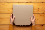 Fototapeta  - Pizza closed carton box in human caucasian hands on natural wooden table flat lay mockup with blank space