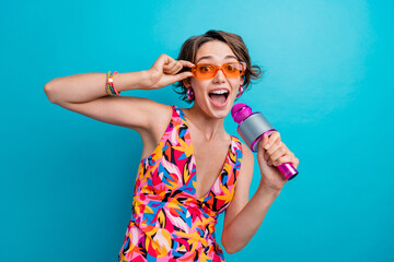 Poster - Portrait of astonished girl with short hairstyle wear colorful dress hold microphone touching sunglass isolated on blue color background