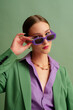 Fashionable beautiful confident woman wearing trendy purple color rectangular sunglasses, suit blazer, office shirt, chain necklace, posing on green background. Studio fashion close up portrait