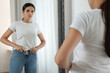 Young woman trying to put on tight jeans near mirror indoors