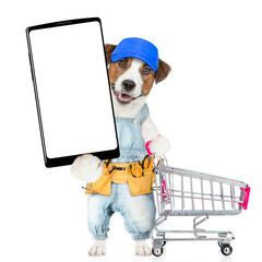 Poster - Funny Jack russell terrier puppy wearing blue cap and denim overalls with tool belt showing big smartphone with white blank screen in it paw and pushing the shopping cart. isolated on white background