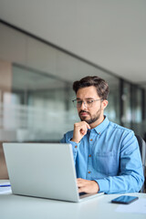 Busy professional business man company employee, young male worker software developer, latin businessman looking at computer thinking on online market analysis using laptop working at office. Vertical