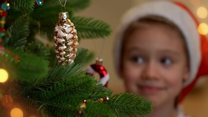 Wall Mural - Close up of happy little girl in red santa hat decorate Christmas tree with golden cone at home. New Year and Christmas