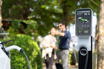 Young couple recharge electric car's battery from charging station in outdoor green city park in springtime. Rechargeable EV car for sustainable environmental friendly urban travel lifestyle.Expedient