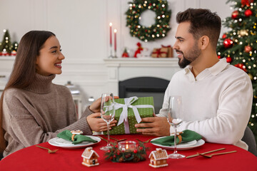 Canvas Print - Happy young man presenting Christmas gift to his girlfriend at table indoors