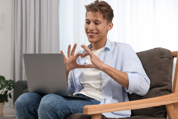 Poster - Happy young man having video chat via laptop and making heart with hands on armchair indoors. Long-distance relationship