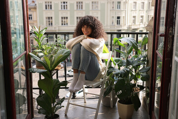 Beautiful young woman relaxing in chair surrounded by green houseplants on balcony