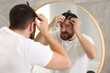 © New Africa - Dandruff problem. Man with comb examining his hair and scalp near mirror indoors