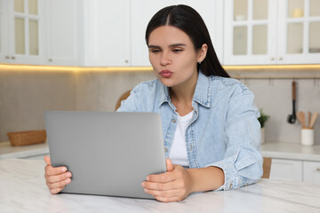 Canvas Print - Young woman having video chat via laptop and sending air kiss at table in kitchen