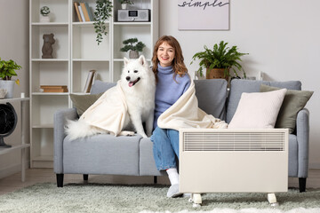 Poster - Young woman with plaid and Samoyed dog on sofa warming near radiator at home