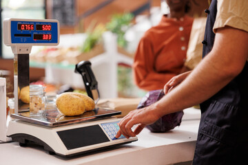 Wall Mural - Diligent seller in a small grocery store measures the weight of organic potatoes using an electronic scale. Caucasian man employed as cashier assisting a customer in weighing locally grown potatoes.