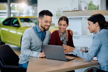 Young happy couple using laptop with car saleswoman in showroom.