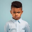 Portrait of an angry little Latin boy with brown hair. Closeup face of a furious Latin American child on a blue background looking at camera. Front view of an outraged irritated kid in a blue shirt.