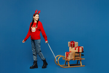 Wall Mural - Full body young Latin woman wearing red Christmas sweater decorative deer horns on head posing carry present box with gift ribbon bow on sled isolated on plain blue background. Happy New Year concept.