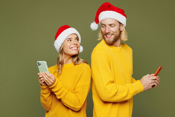 Merry fun young couple two friends man woman wearing sweater Santa hat posing hold use mobile cell phone look to each other isolated on plain green background Happy New Year Christmas holiday concept