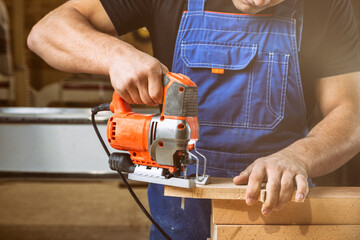 Wall Mural - Close up of experienced carpenter in work clothes and small buiness owner  carpenter saw and processes the edges of a wooden bar with a jig saw  in a workshop