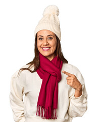 Wall Mural - Middle-aged woman in cozy Christmas winter wear in studio smiling and pointing aside, showing something at blank space.