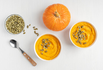 Wall Mural - Top view of homemade creamy pumpkin soup made of ripe squash mashed or pureed with dairy cream and broth decorated with seeds served in bowls on white wooden background with spoon for healthy dinner