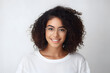 Close up studio shot of beautiful young mixed race woman model with curly dark hair looking at camera with charming cute smile while posing against white blank copy space wall for your content