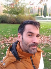 Selfie portrait of Caucasian man looking at camera with short dark hair in brown and white coat suspicious with raised eyebrow and looking at camera in a park during autumn winter