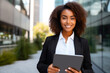 © MVProductions - Black businesswoman smiling and holding tablet, with office building in the background