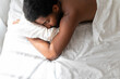 Young African American man sleeping soundly in his comfortable bed at home, lying on stomach, hugging a pillow