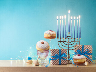 Wall Mural - Jewish holiday Hanukkah creative composition with menorah, traditional donuts and gift box on wooden table over blue background