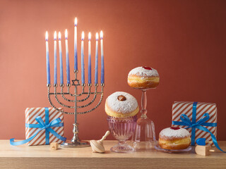 Wall Mural - Jewish holiday Hanukkah creative composition with menorah, traditional donuts and gift box on wooden table over modern background