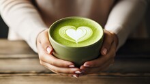Woman's Hands Holding Green Cup Of Matcha Latte With Heart Shape Latte Art On Wooden Background