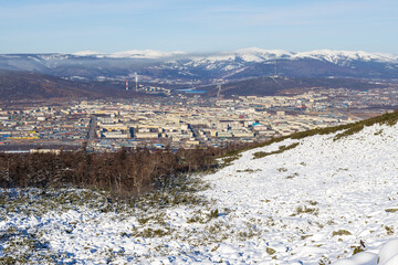 Wall Mural - View from the mountain to the city of Magadan. In the foreground is a snow-covered hillside. In the distance there is a city and snow-capped mountains. Northern nature. Magadan, Magadan region, Russia