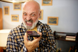 Fototapeta  - Senior man in checkered shirt yelling at smartphone. Angry furious bald bearded man recording voice message, expressing rage and displeasure, shouting with opened mouth, scolding someone